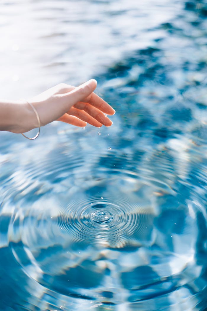 A woman's hand gently creates ripples on the surface of a blue swimming pool under bright sunlight.